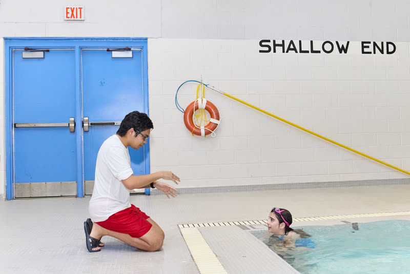 A lifeguard kneels at the shallow end beside a swimmer, reaching assist and buoy within reach.