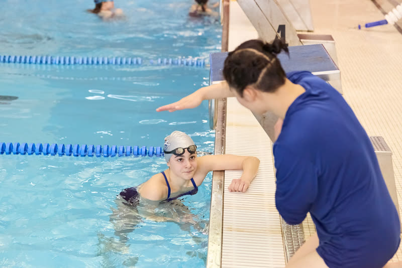 A coach on the deck gives stroke feedback to a swimmer resting at the wall. Teens & Adults program.
